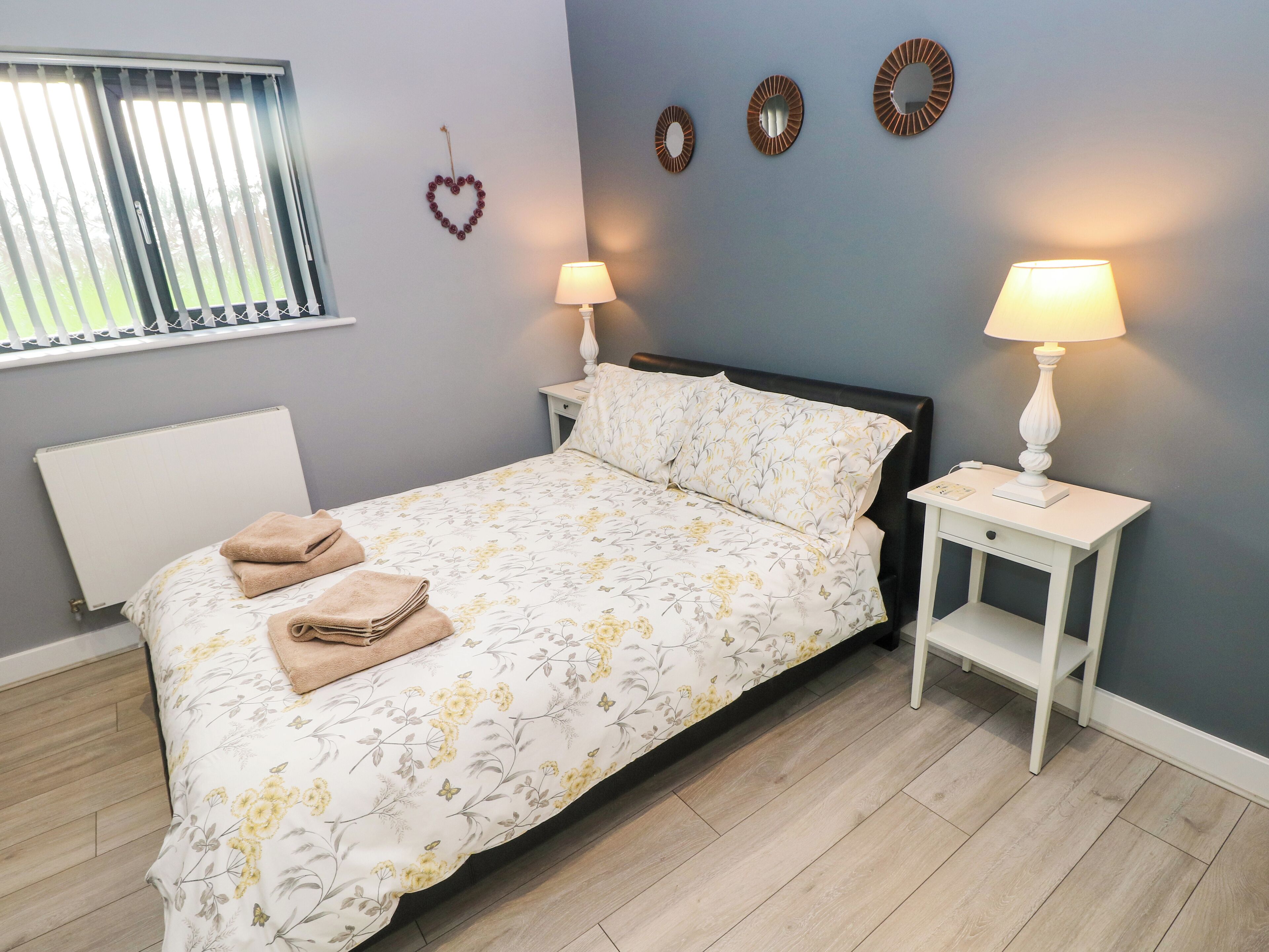 Un lit bien fait, avec une literie à motifs floraux, deux tables de chevet avec des lampes, un radiateur et une fenêtre avec des stores.