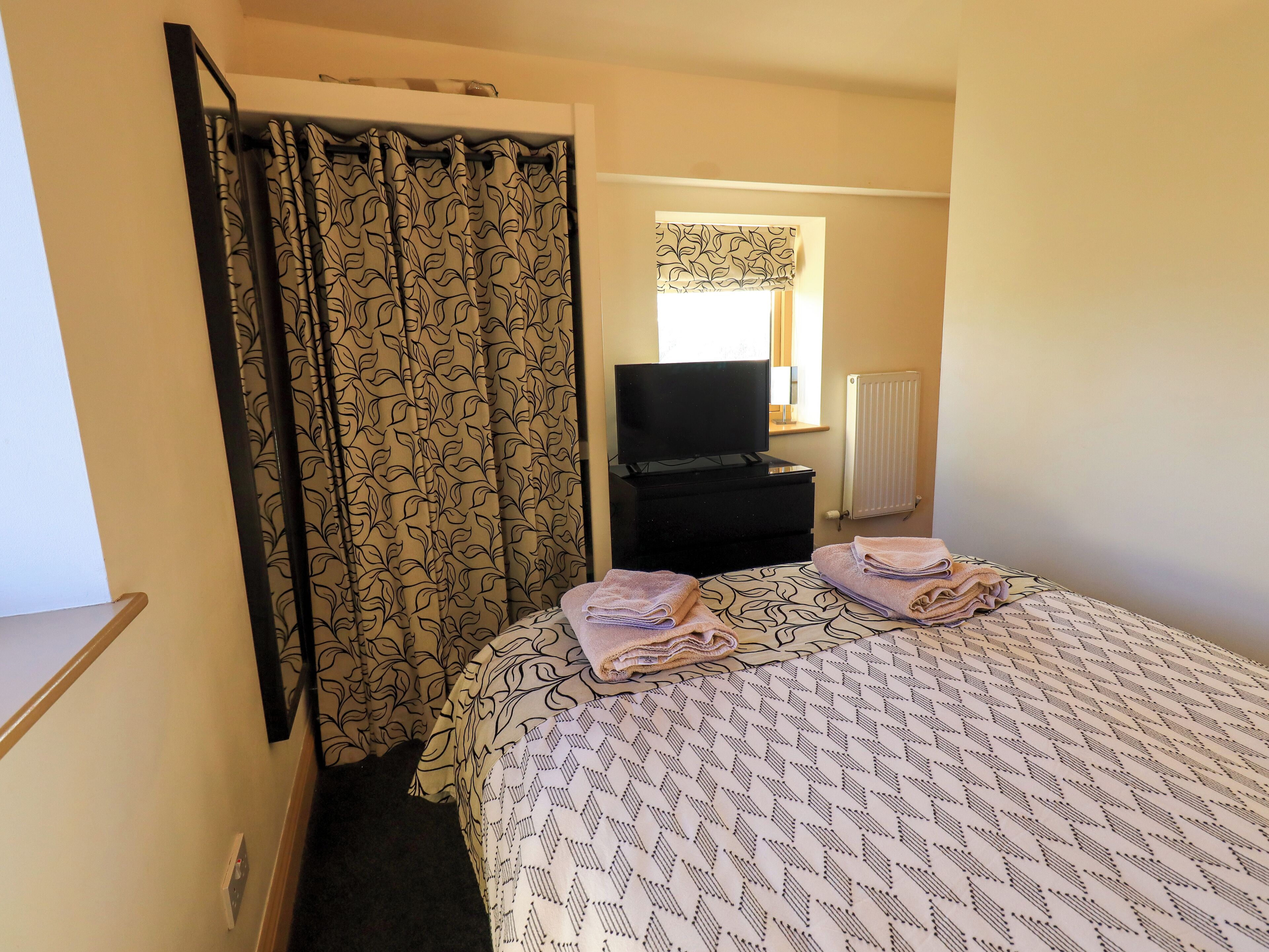 Una habitación de hotel con una cama, una silla negra, un televisor y una ventana con cortinas.