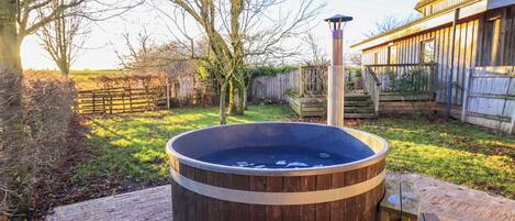 A wooden hot tub with a metal heater, surrounded by a wooden deck, a wooden fence, and a wooden building in the background.