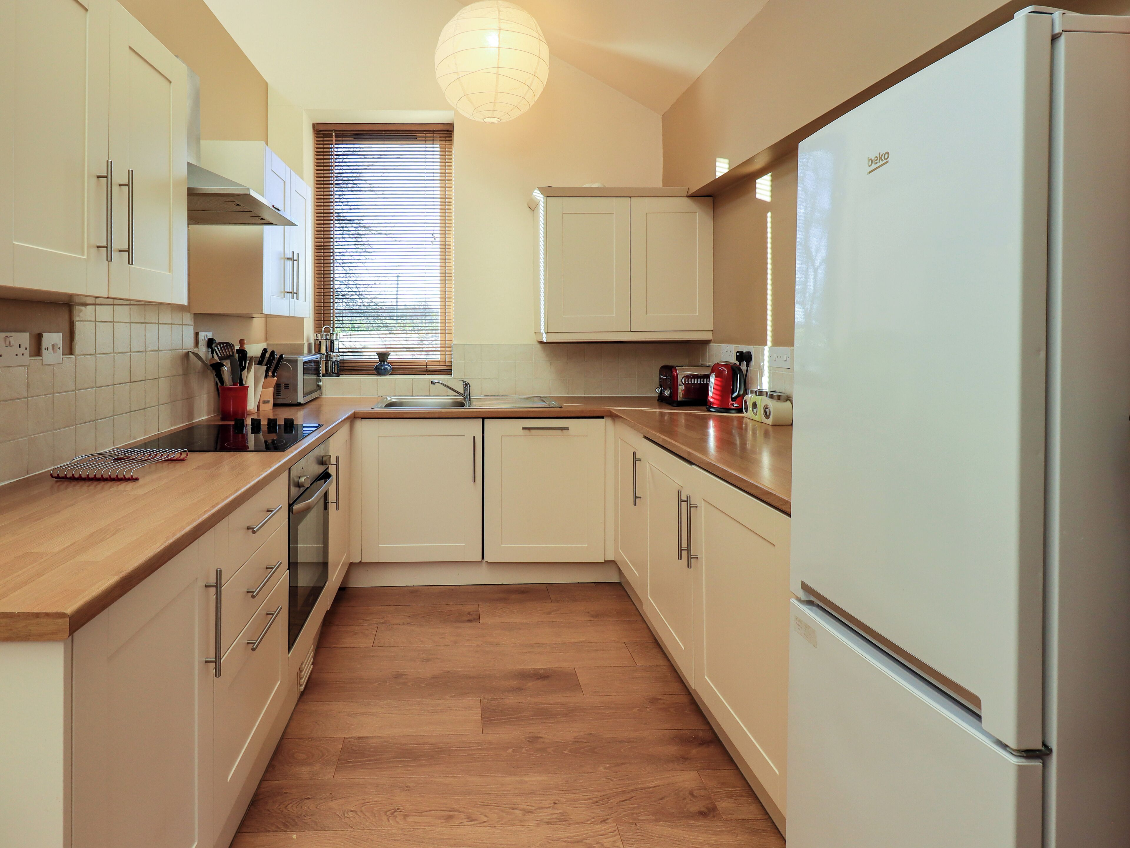 A modern kitchen with wooden flooring, white cabinets, a stainless steel refrigerator, and a window with blinds.