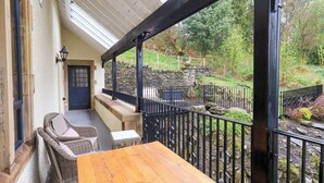 A wooden table and wicker chairs on a covered porch with a stone wall and metal railing in the background. - Deer Wood At Applethwaite Hall (Windermere)