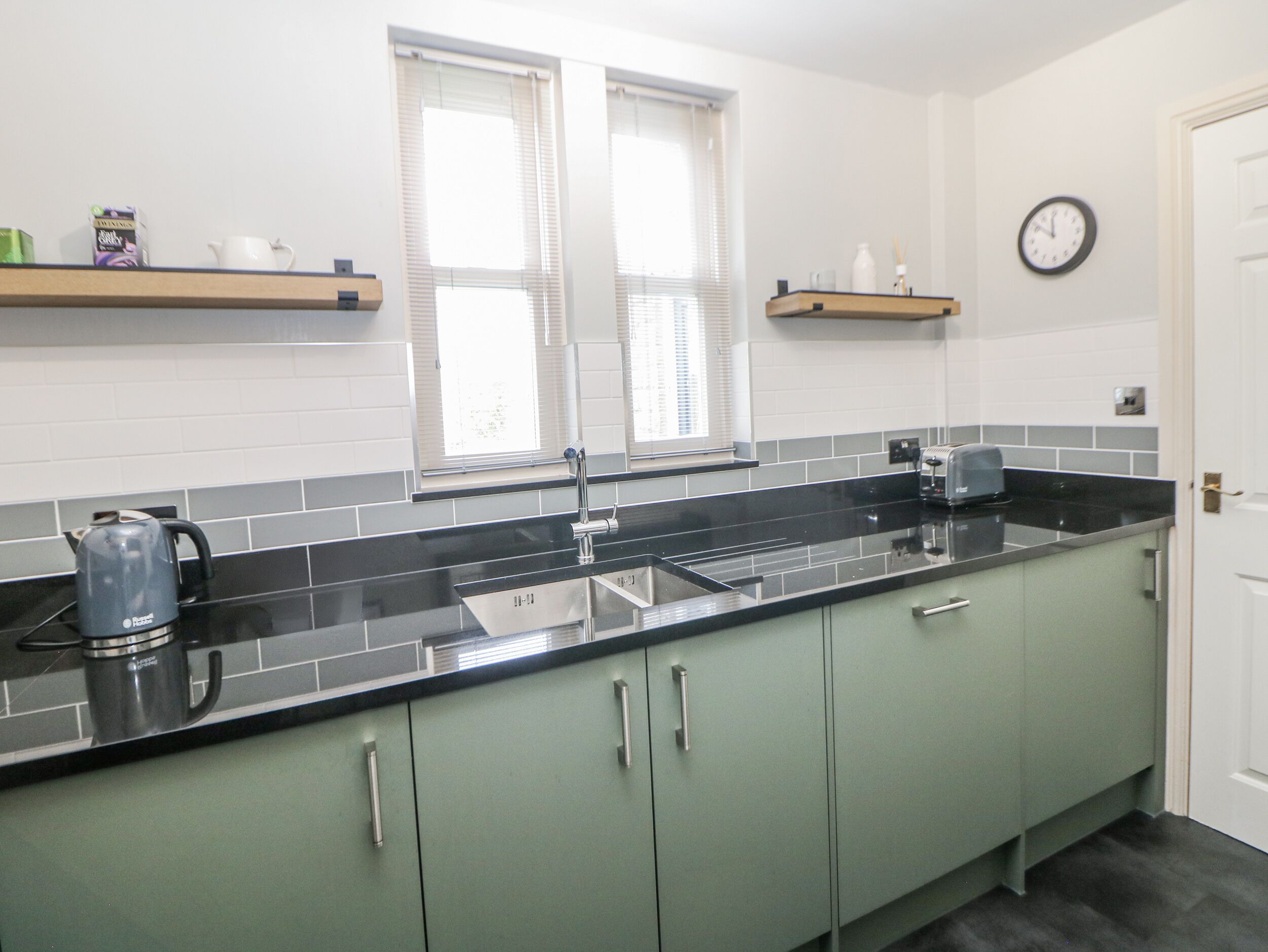 A kitchen with a black countertop, stainless steel sink, and green cabinets. There are two windows with blinds, a clock on the wall, and a shelf with kitchen items.