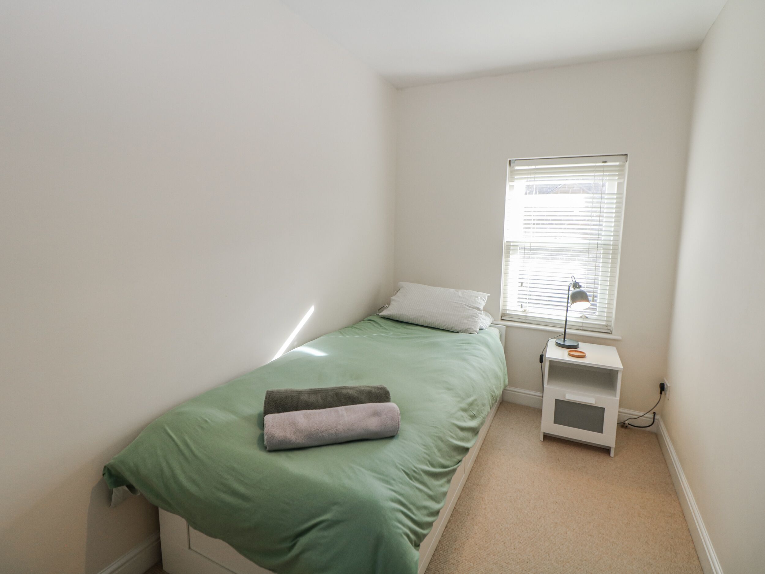 A small, neatly arranged bedroom with a single bed, a bedside table with a lamp, and a window with blinds.