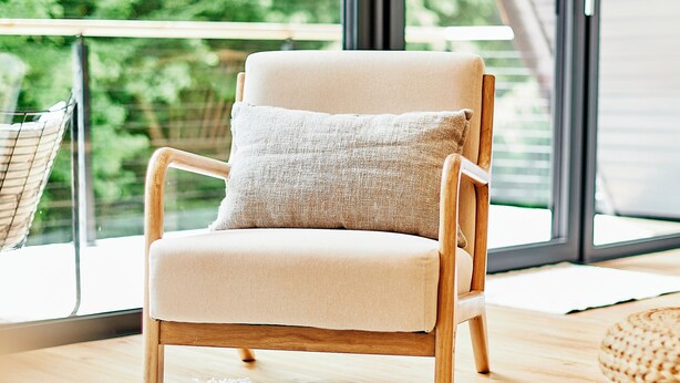 A modern chair with a cushion, a white rug, and a view of greenery through a large window. - HOUSE28 - Modern A-Frame in the woods (Grattersdorf)