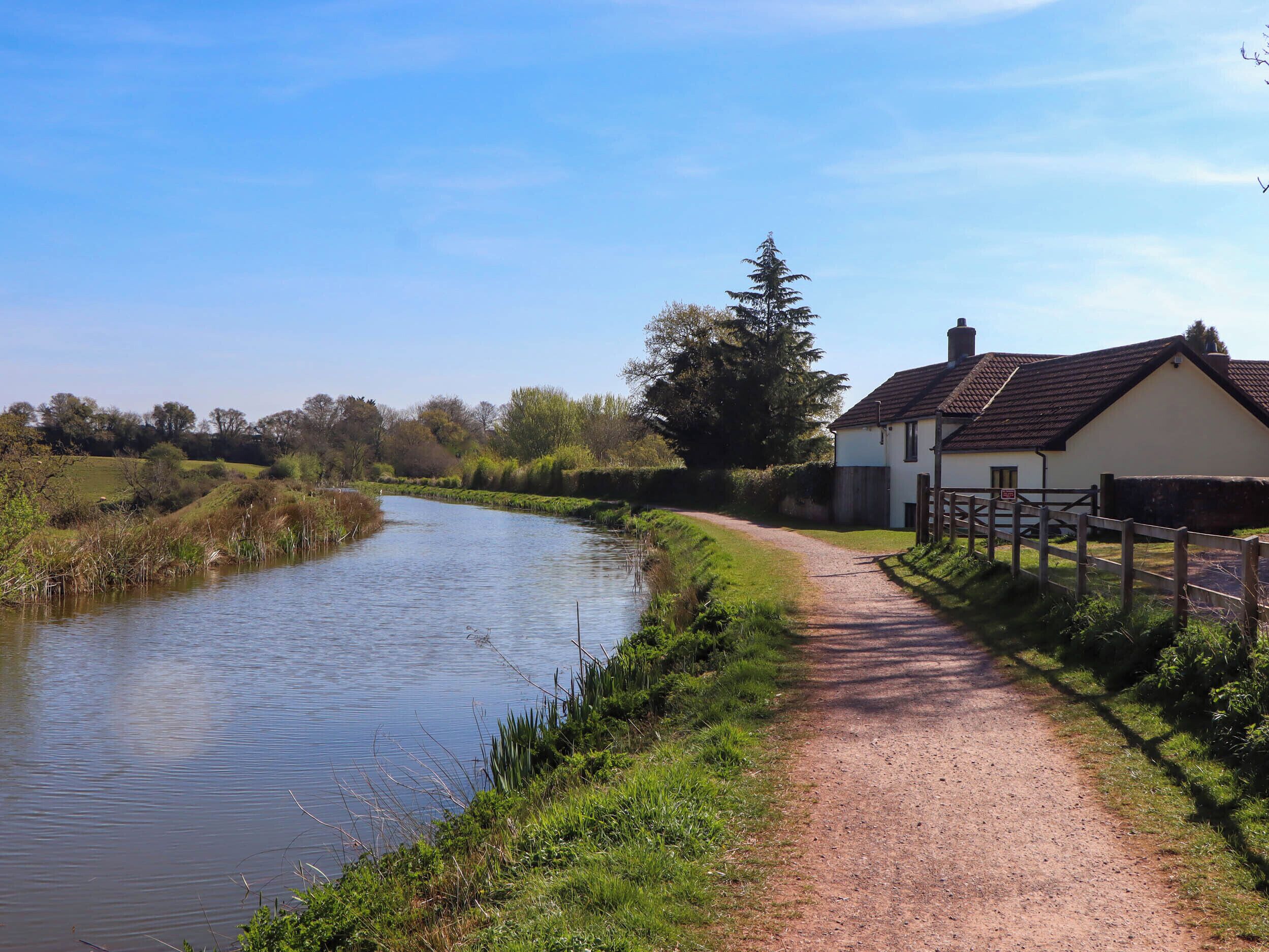 A rural pathway beside a calm river, leading to a white house with a dark roof.
