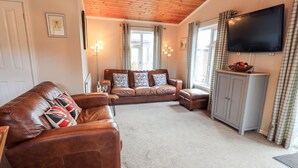 A living room with a brown leather sofa, a matching ottoman, and a two-seater sofa with patterned cushions. A flat-screen TV is mounted on the wall, and a cabinet with a fruit bowl is visible. - Brightwater Lodge (Windermere)