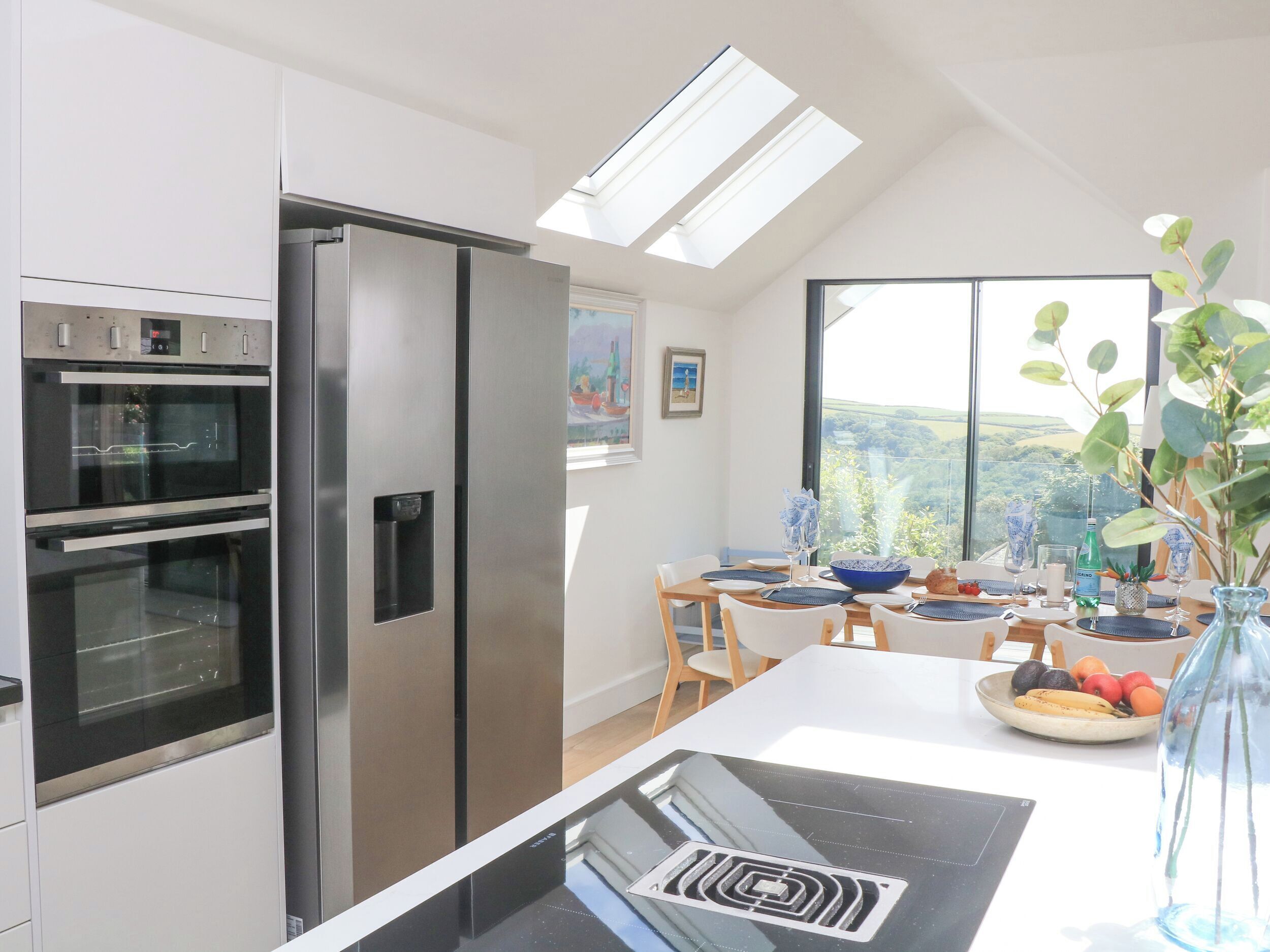 A modern kitchen with a built-in oven, a stainless steel refrigerator, and a dining area with a large window offering a scenic view.