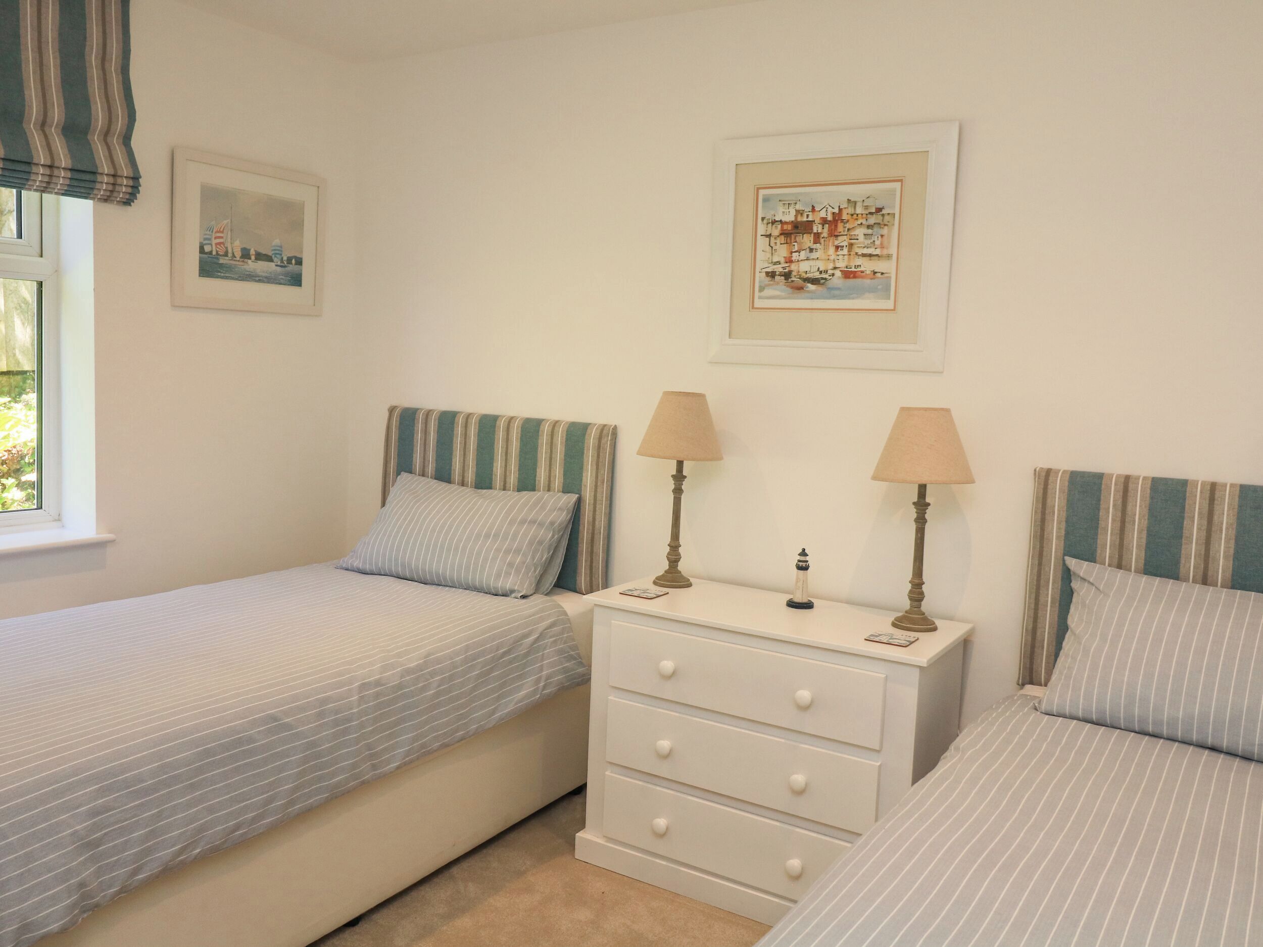 A bedroom with two single beds, a bedside table with lamps, a dresser, and two framed pictures on the wall.