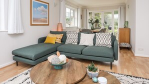 A modern living room with a sectional sofa, a round wooden coffee table, and a view of the garden through large windows. - The Palms (Pwllheli)
