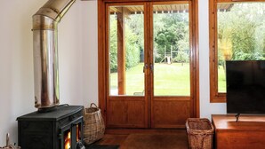 Ein gemütliches Wohnzimmer mit Kaminofen, Fernseher und Blick auf einen grünen Garten durch große Holztüren.