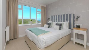 A bedroom with a large bed, a bedside table, a window with curtains, and a view of the landscape. - Rowena (Helston)
