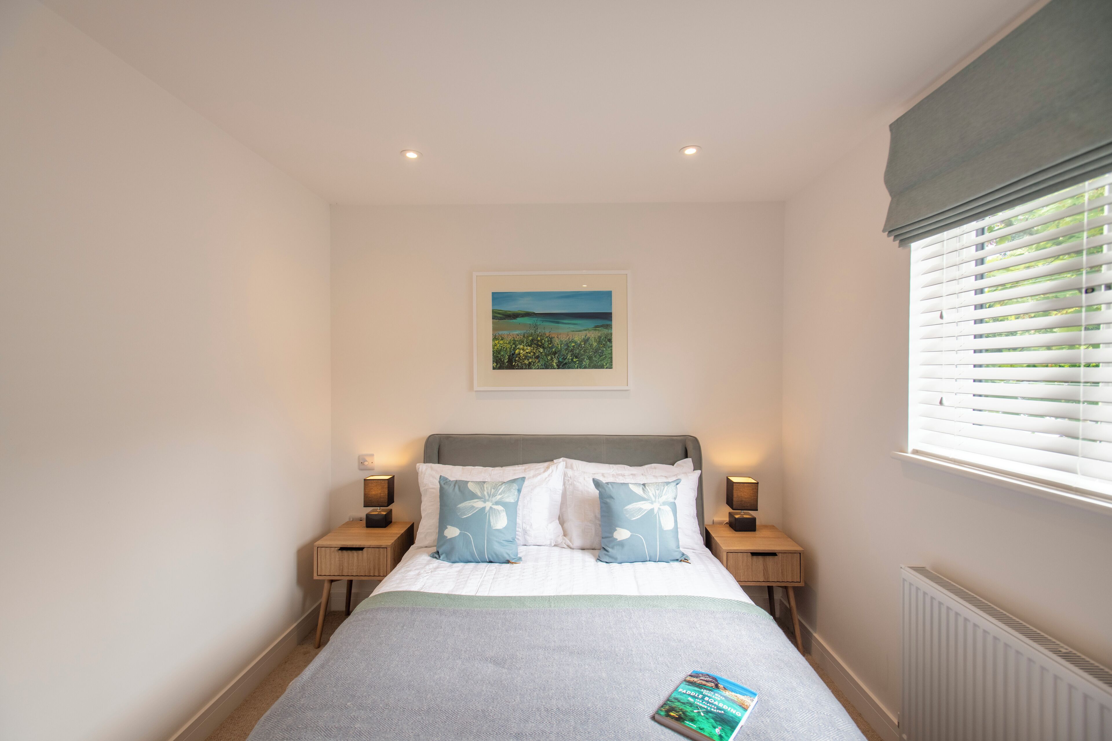 A bedroom with a bed, two bedside tables, a nightstand, a framed picture on the wall, and a window with blinds.