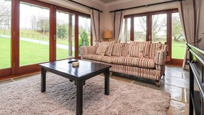 A spacious living room with a striped sofa, a coffee table, and large windows offering a view of a grassy area. - The Barn (Knaresborough)