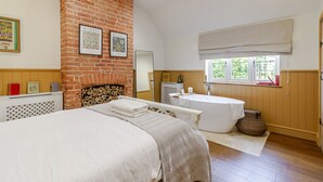 Un dormitorio con chimenea de ladrillo, una cama grande, una mesita de noche, un espejo, una ventana con vistas y un baño con bañera y lavamanos.