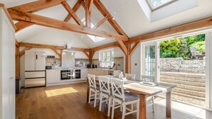 Una cocina amplia con vigas de madera, un comedor con sillas blancas y vistas a una escalera de piedra que da al exterior.