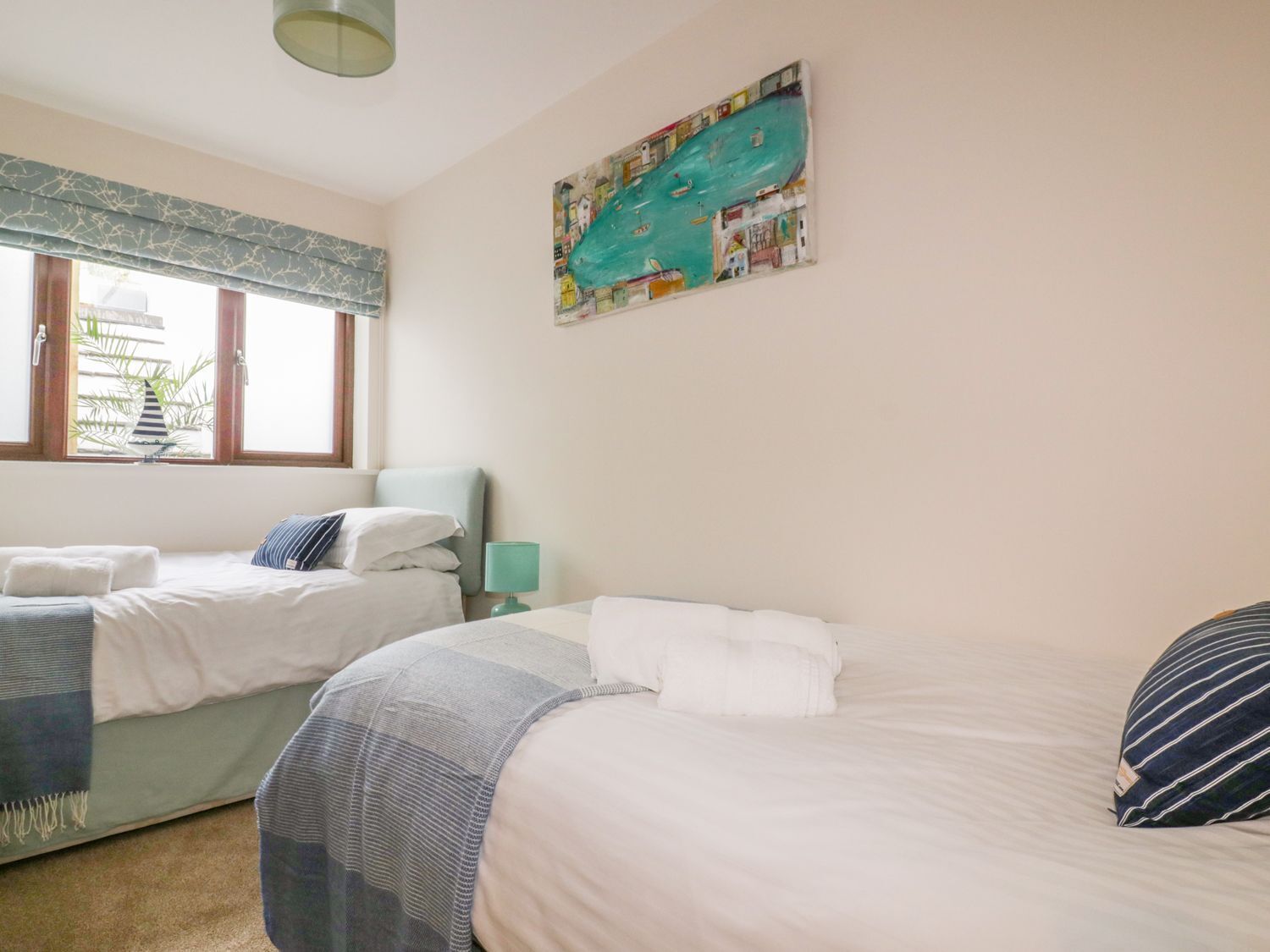 Quarto de hotel com duas camas, uma janela com vista para palmeiras, um quadro na parede e uma luminária na mesinha de cabeceira.