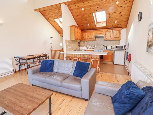 Un accogliente soggiorno con un divano grigio e un tavolino in legno, una cucina con mobili in legno e un frigorifero bianco, e una sala da pranzo con tavolo e sedie.
