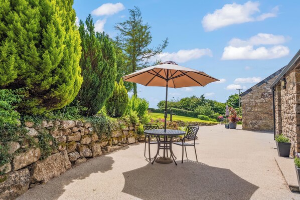 A patio with a table and chairs under an umbrella, surrounded by stone walls and greenery.