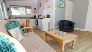A cozy living room with a sofa, a coffee table, and a black armchair. A kitchen with a microwave and a refrigerator is visible in the background. - Chalet 3 Wipeout (Hayle)