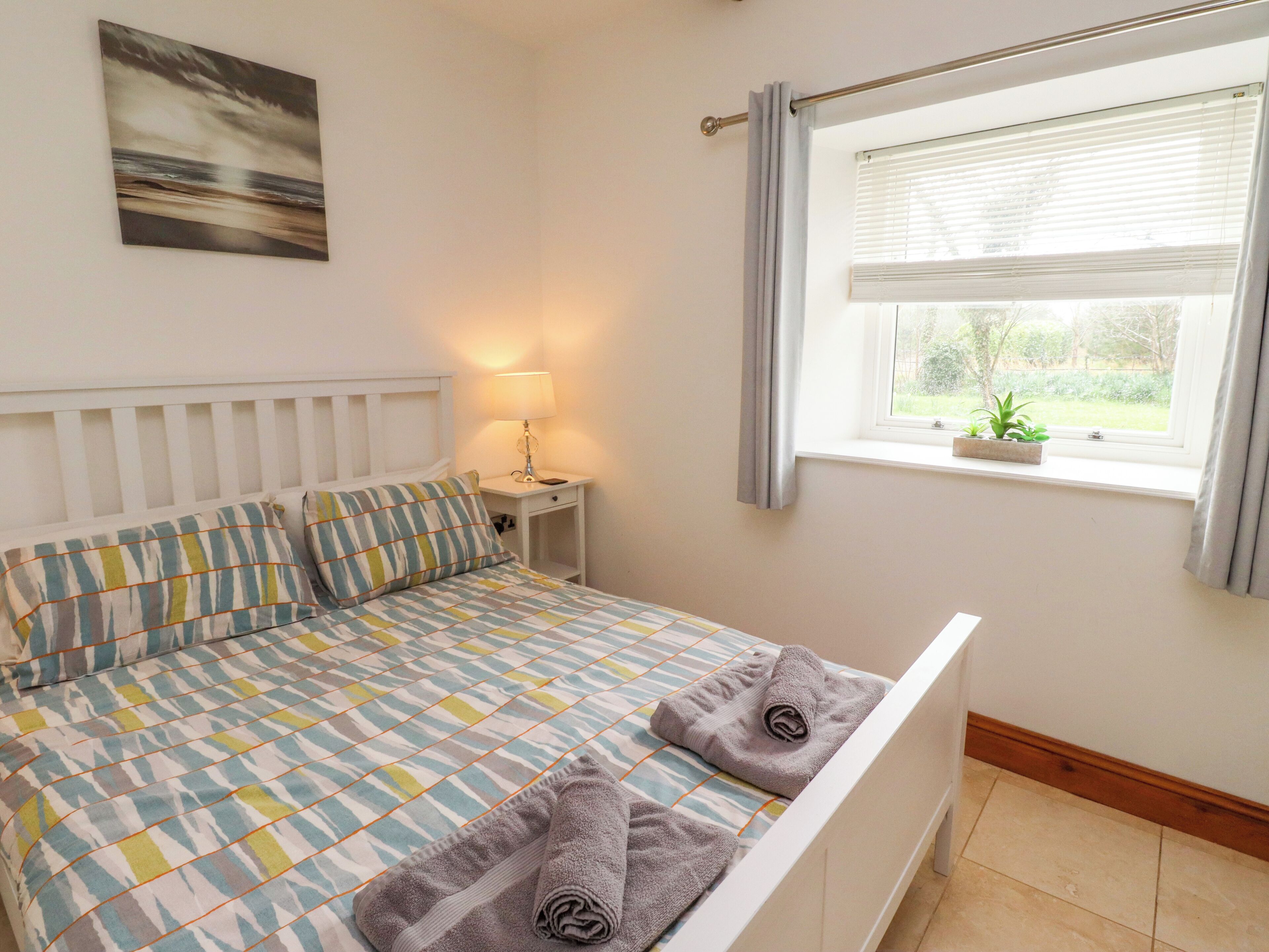 A neatly made bed with striped pillows, a bedside lamp, a window with curtains, and a small plant on the windowsill.