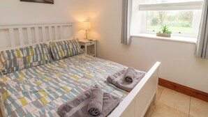 Una cama bien tendida con almohadas a rayas, una lámpara de noche, una ventana con cortinas y una pequeña planta en el alféizar.