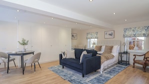 Een moderne woonkamer met een donkerblauwe bank, een eettafel met stoelen, een groot raam met gordijnen en een bed met hoofdeinde.