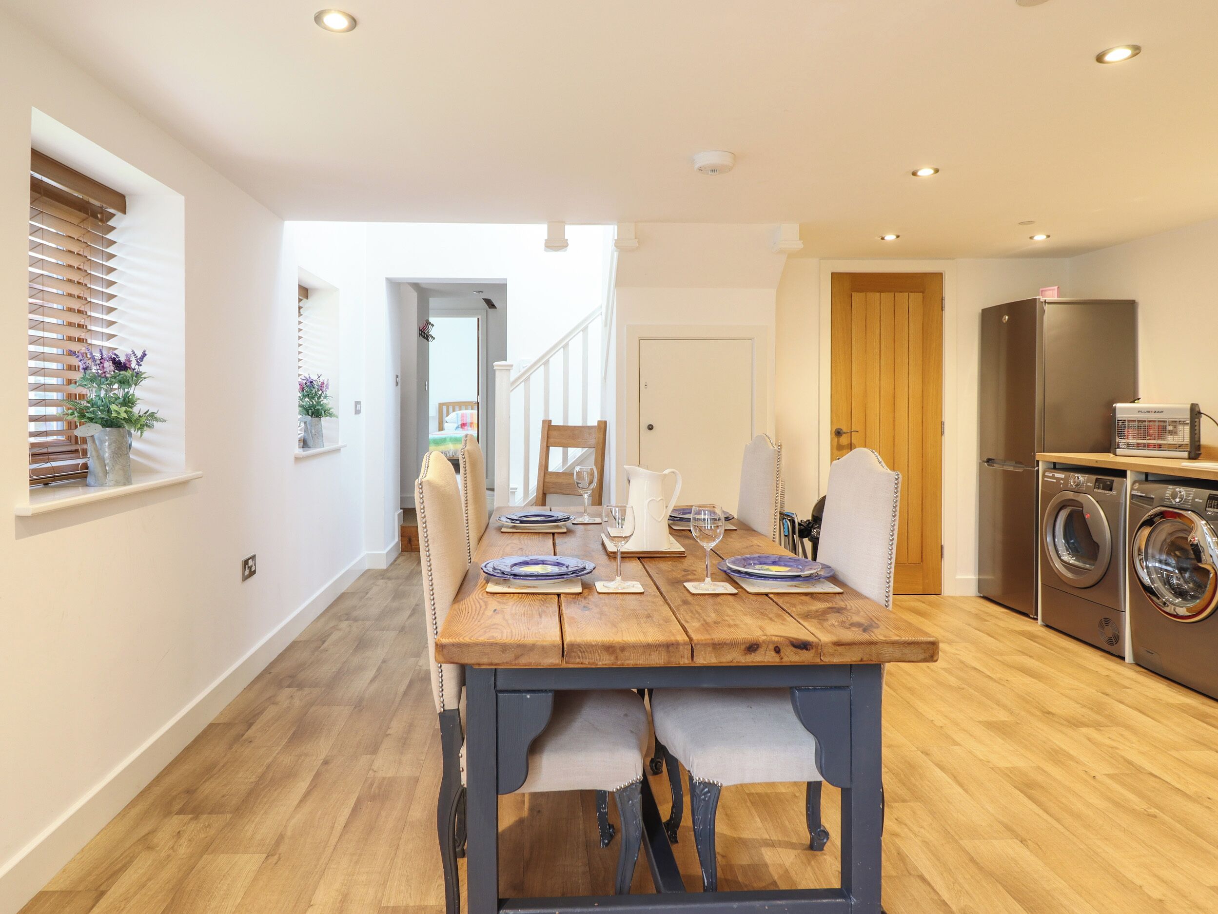 Un coin repas avec une table en bois dressée pour quatre personnes, une cuisine équipée d’un lave-linge et un escalier menant à une autre pièce.