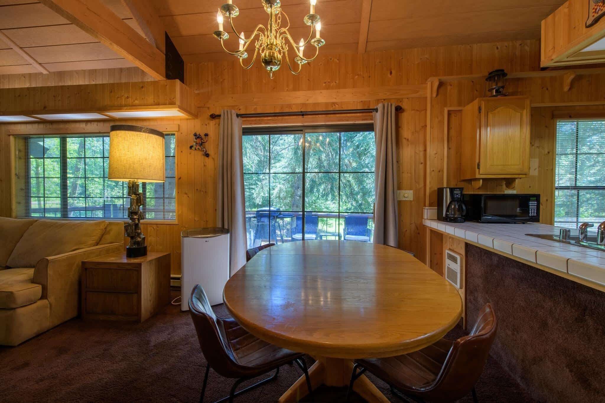 Un intérieur en bois avec une table de salle à manger ronde, des chaises, un canapé, une lampe, un lustre et un coin cuisine équipé d’un four à micro-ondes, ainsi que de grandes fenêtres avec des rideaux.