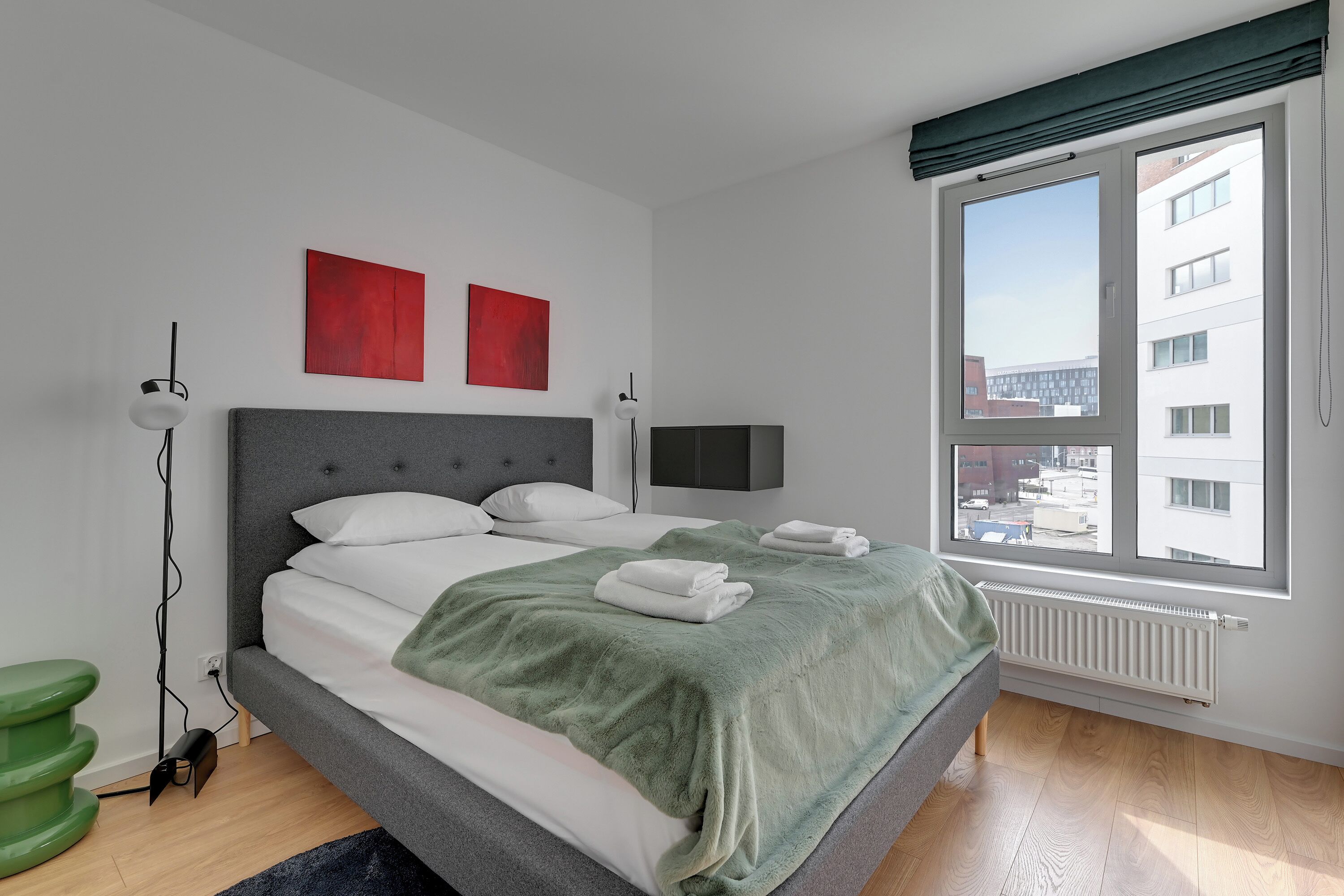 Ein modernes Schlafzimmer mit einem Bett, einer grünen Decke, einem roten Kunstwerk, einem Fernseher und einem Fenster mit Blick auf die Stadt.