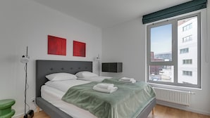 A modern bedroom with a bed, a green blanket, a red artwork, a TV, and a window with a city view. - RentPlanet - Apartamenty Żurawie (Gdansk)