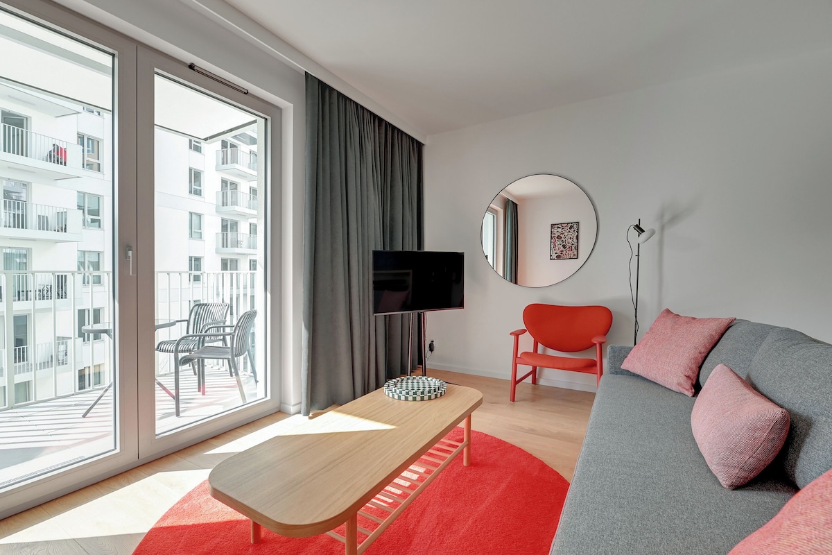 Ein modernes Wohnzimmer mit einem großen Schiebeglasfenster, einem Holztisch, einem grauen Sofa mit rosafarbenen Kissen, einem roten Sessel und einem Flachbildfernseher.