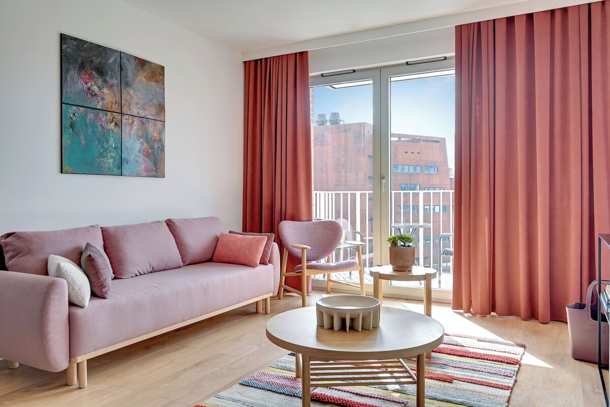 Ein modernes Wohnzimmer mit einem rosafarbenen Sofa, einem runden Couchtisch und einem Sessel. Ein Balkon mit roten Vorhängen und Blick auf die Stadt.