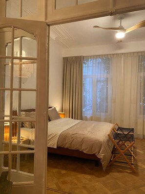 Relax in this serene bedroom with a large bed against a window, a ceiling fan above, and a comfortable chair nearby.