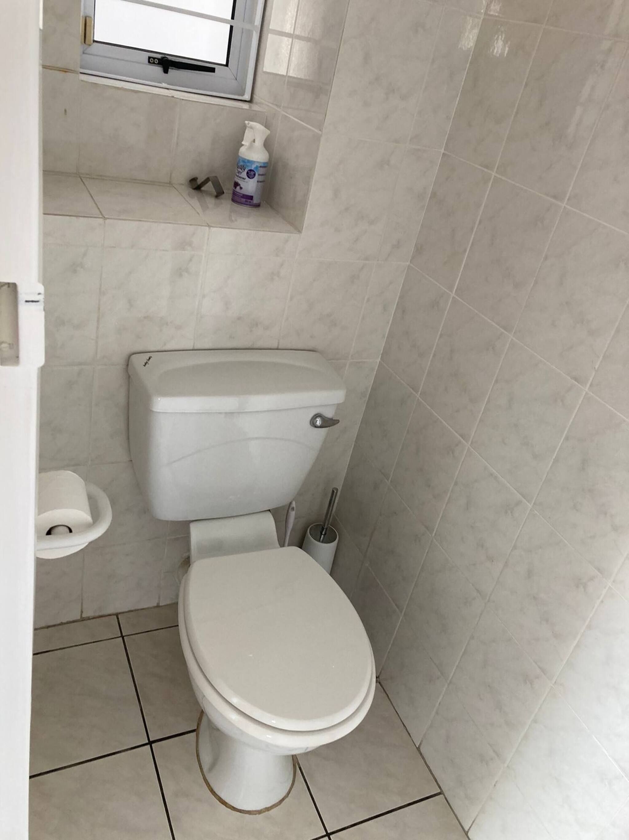 A small, clean bathroom with a toilet, a small shelf with a bottle, and a window above the shelf.