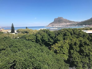 Un pueblo costero con abundante vegetación en primer plano, una colina destacada con una construcción similar a una fortaleza, y una vista despejada del mar.