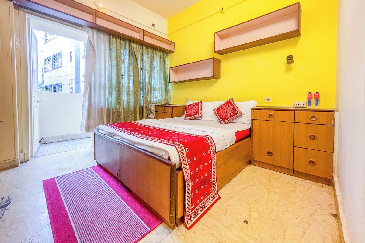A hotel room with a bed, a wooden nightstand, a small table, a window with curtains, and a yellow wall.