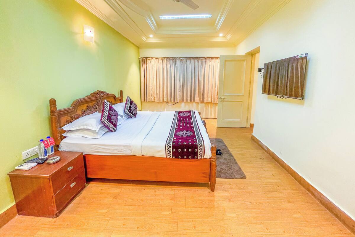 A bedroom with a wooden bed, a nightstand, a desk, a ceiling fan, a window with curtains, and a door.