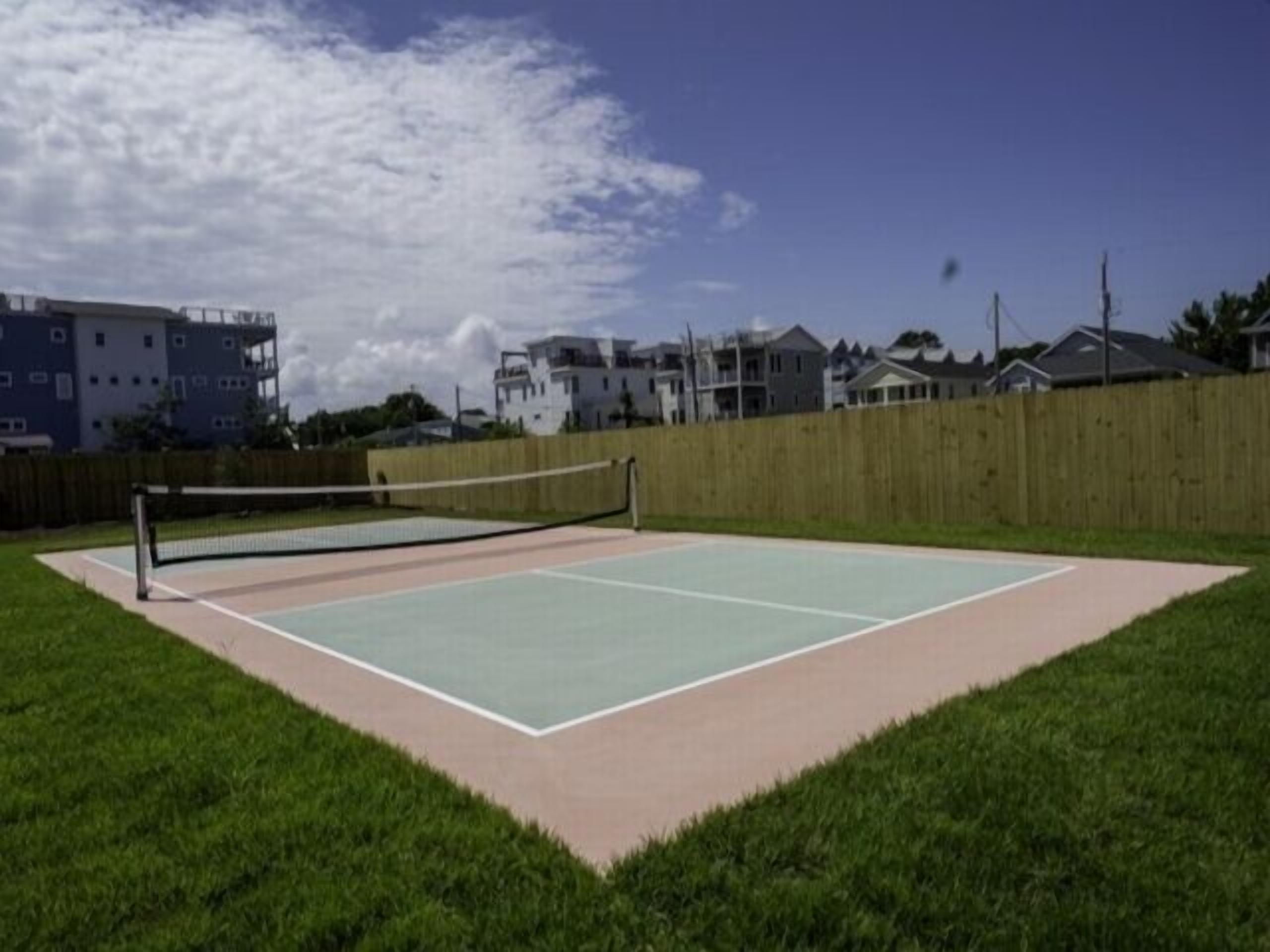 An outdoor tennis court with a wooden fence and houses in the background.