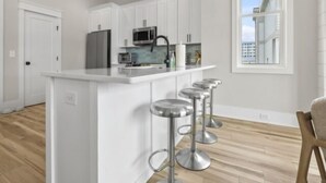A modern kitchen with white cabinetry, stainless steel appliances, and a central island with bar stools. - A Treasure To Sea 4 Bedroom Villa (Carolina Beach)