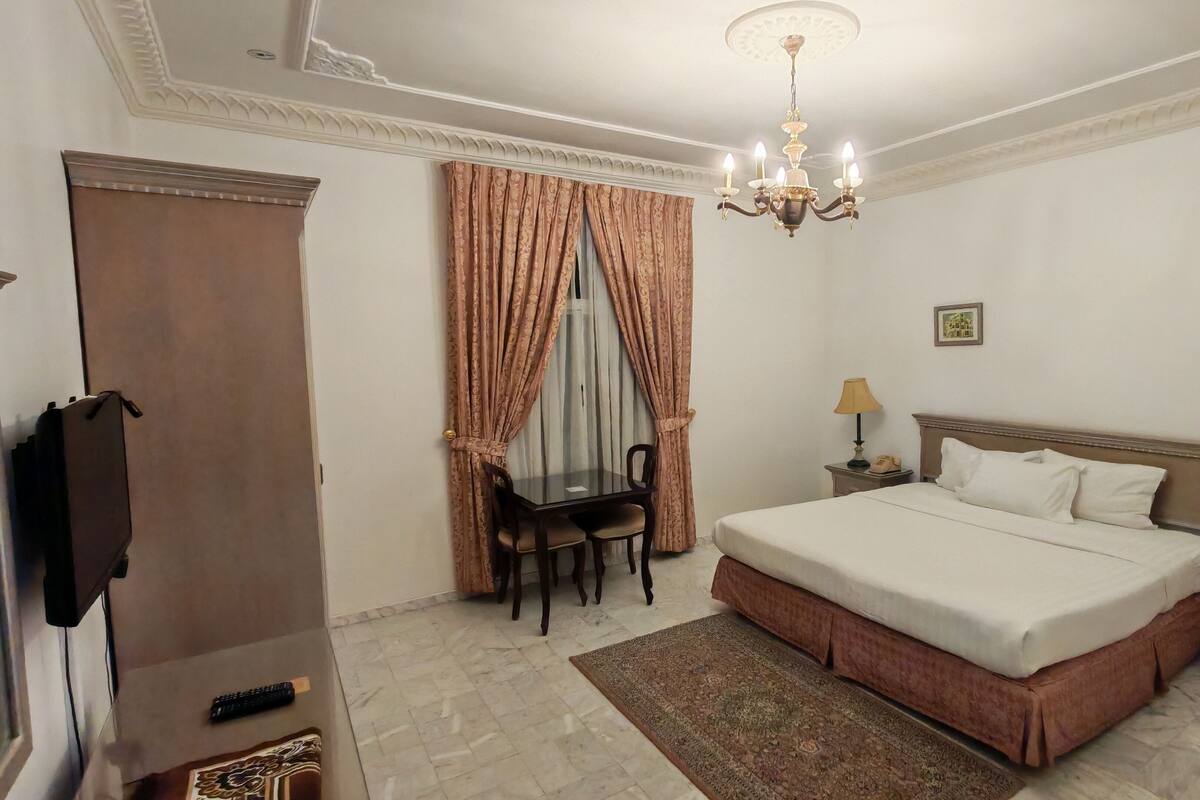 A hotel room with a large bed, a desk with a chair, a chandelier, and two bedside lamps.