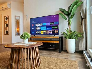 Ein Wohnzimmer mit Fernseher, der Streaming-Dienste anzeigt, einem Holztisch und zwei Blumentöpfen.