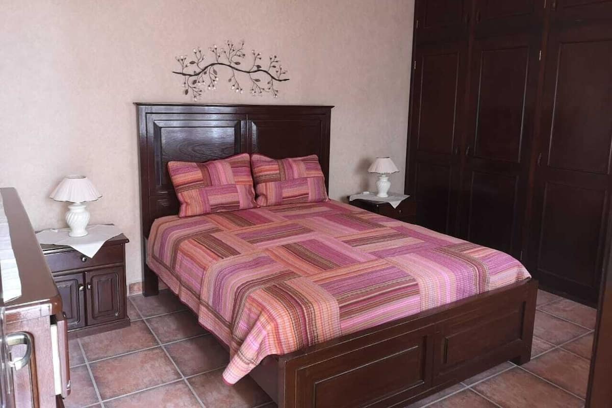 Quarto com cama de madeira, duas mesas de cabeceira com abajures e um elemento decorativo acima da cama.