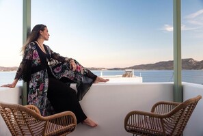 A woman sitting on a balcony with wicker chairs, overlooking a body of water and distant hills. - Eclipse Tales (Milos)