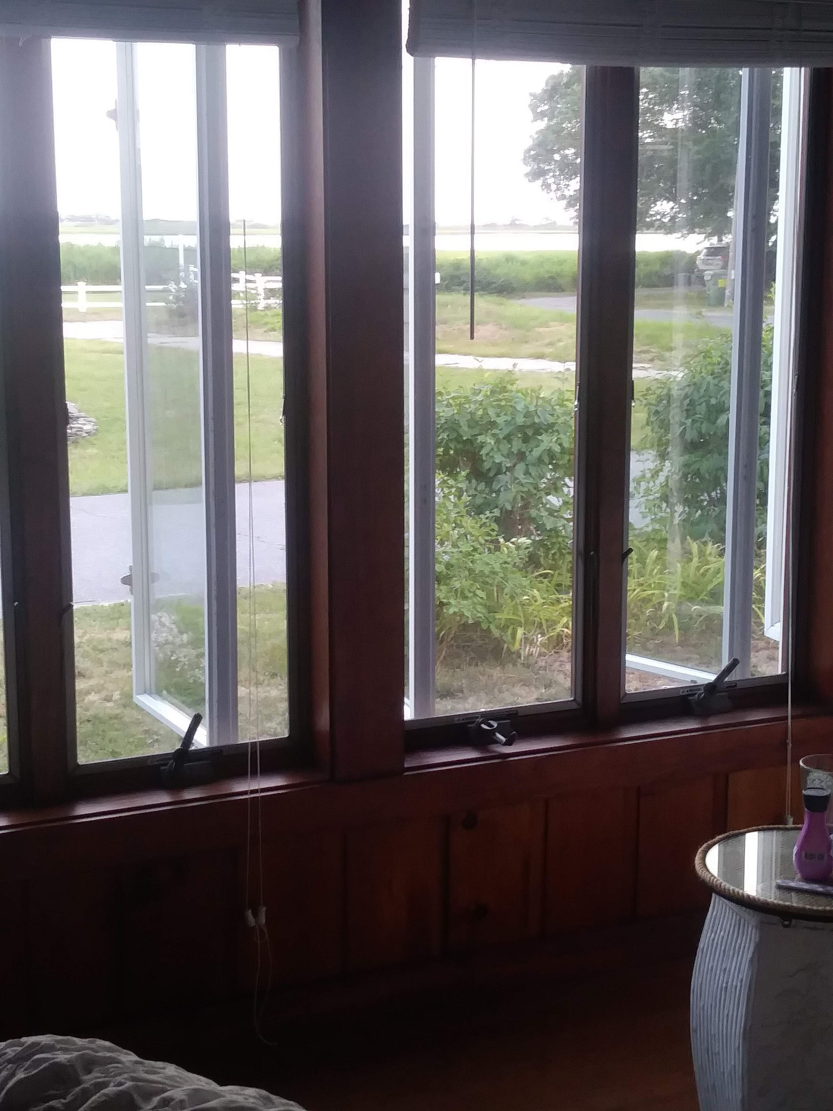 A room with large windows overlooking a garden and a road. There is a small table with a vase and a bottle on it.