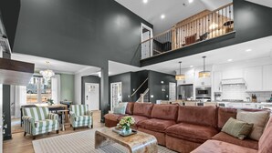 A spacious living area with a large sofa, striped armchairs, and a coffee table. A staircase leads to a balcony with a railing. - 6BR w/ Golf Sim Putting Green Hot Tub Sauna Games (Pinehurst)
