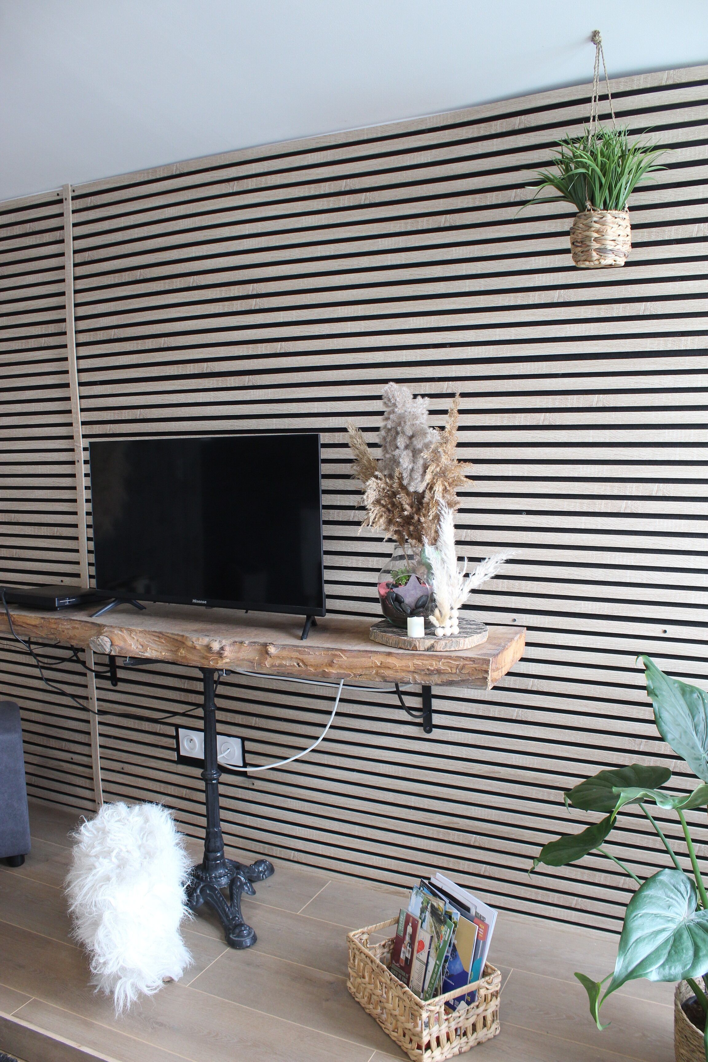 Un salon moderne avec un téléviseur à écran plat, une table basse en bois, un lampadaire noir, un tapis blanc et moelleux, un panier en osier rempli de magazines et une plante suspendue.