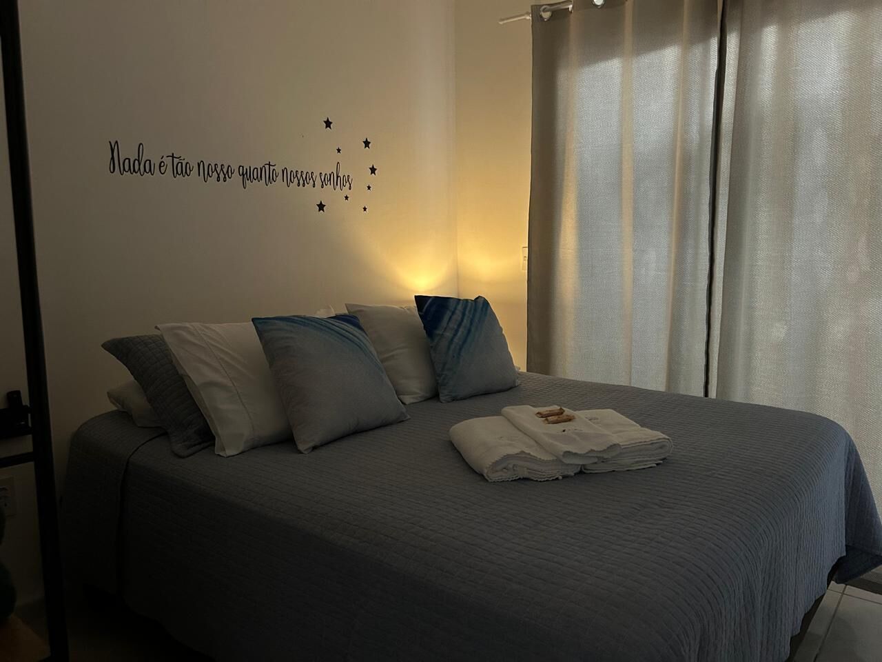 A neatly made bed with a grey bedspread and pillows, a folded towel on the bed, and a wall with a star decal and text.