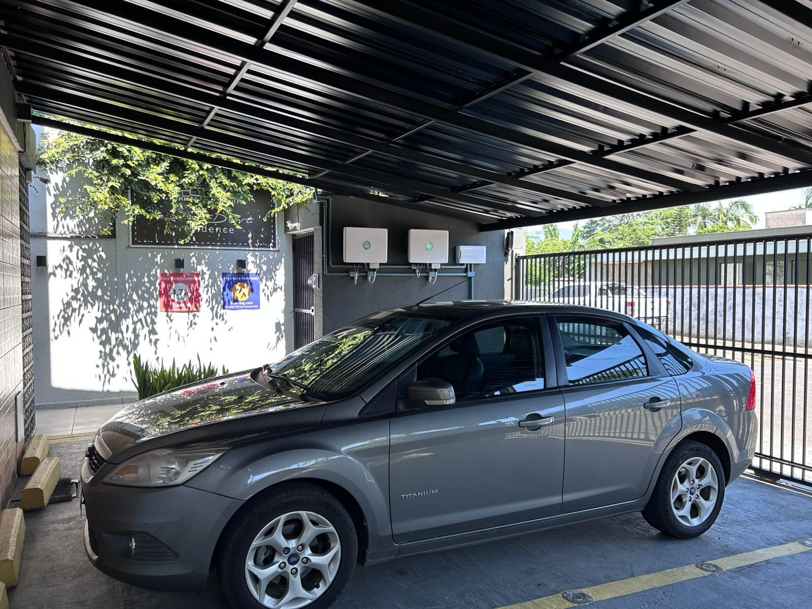 Um carro estacionado sob uma cobertura com portão e um edifício com uma placa ao fundo.