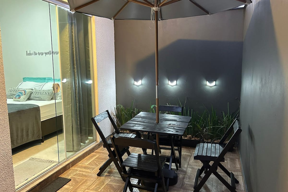 A patio with a table and chairs, a glass wall with a bed and pillows, and wall-mounted lights.
