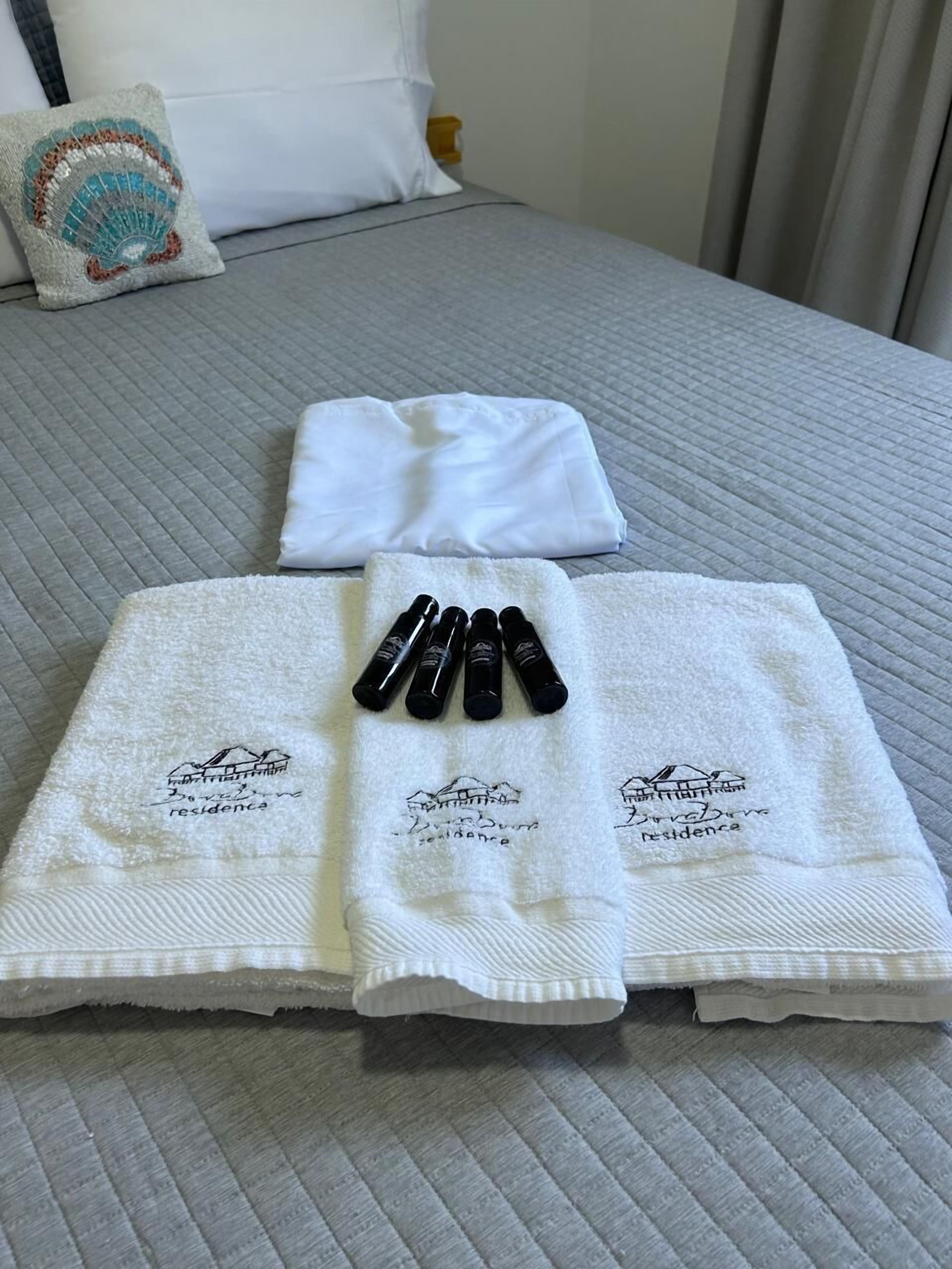 Cama bem arrumada com colcha cinza acolchoada, duas toalhas brancas com texto bordado e quatro garrafas pretas organizadas sobre uma toalha.